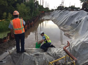 A worker collects a sample of construction site stormwater runoff in a plastic-lined settling pond while another looks on.