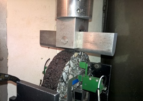 A sample disk of asphalt stands vertically in testing equipment to be compressed from one edge to the other.