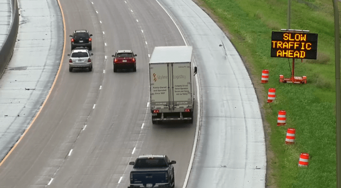 An aerial view of a multilane highway showing light traffic in two visible lanes. A variable message sign alongside the highway displays the warning “Slow Traffic Ahead.”