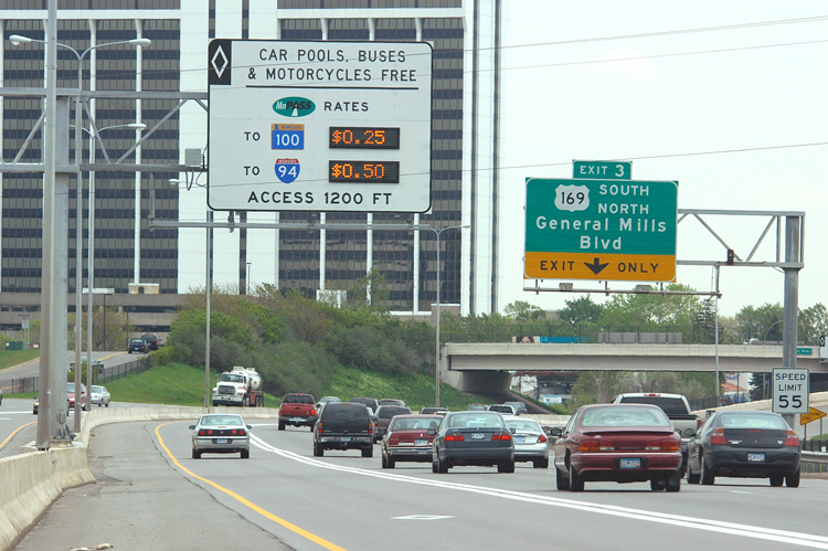 A MnPASS lane on Interstate 394 at the General Mills Boulevard exit. The express lane is closest to the highway median, indicated by a white diamond-shaped marker on the pavement and separated from three other traffic lanes by a solid white line. A highway sign above the lane indicates the fees for lane use.