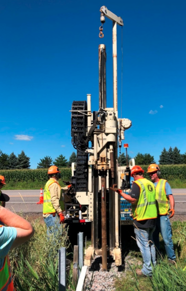 Three workers in safety vests and hard hats stand near a tall boring machine taking a core sample of the ditch check filter.