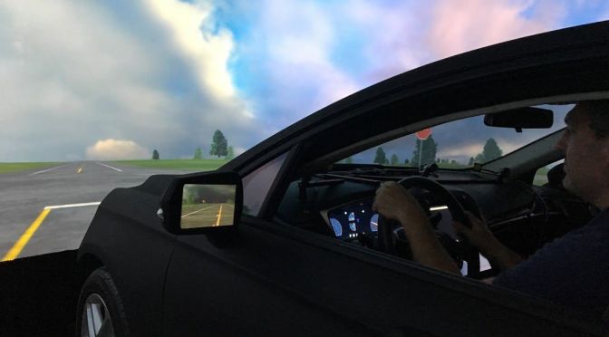 The driver side of the driving simulator. The driver is behind the wheel facing a realistic image of a wide-open rural intersection.