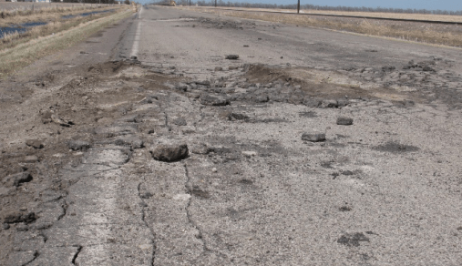 A rural asphalt road shows badly deteriorated areas, with cracks and holes in the asphalt.