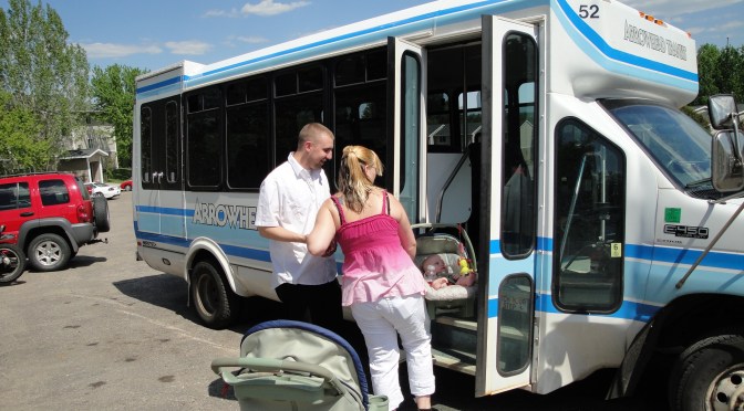 A man and woman stand outside a white commercial bus. The doors of the bus are open and the woman prepares to board with her infant in a carrier.
