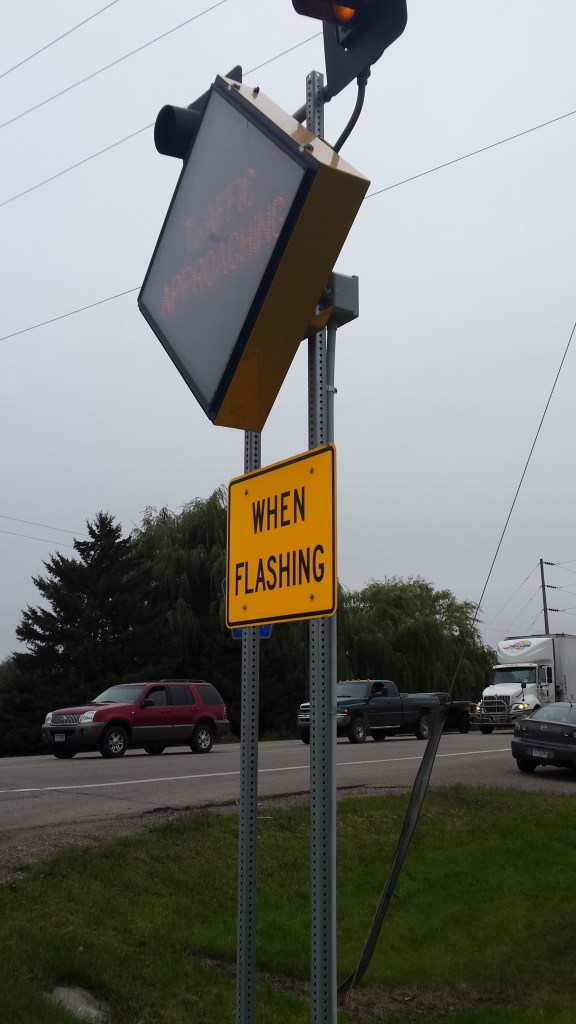 A diamond-shaped RICWS installed alongside a rural highway. A yellow light at the top of the sign flashes when an oncoming vehicle approaches the intersection and traffic is present on the major roadway.