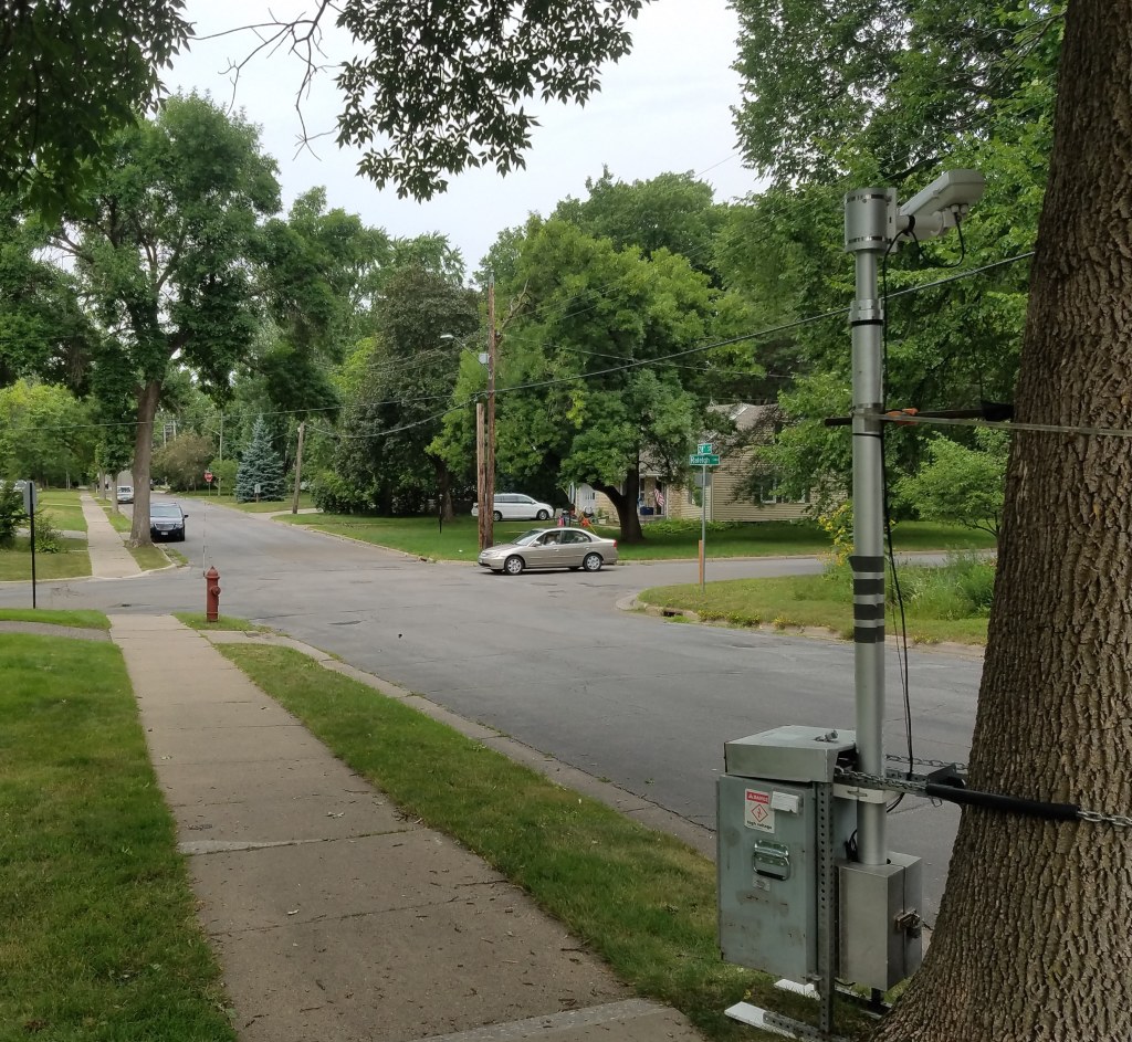 A tall video camera with enclosed power source is attached to a tree near the sidewalk along a two-lane city street. 