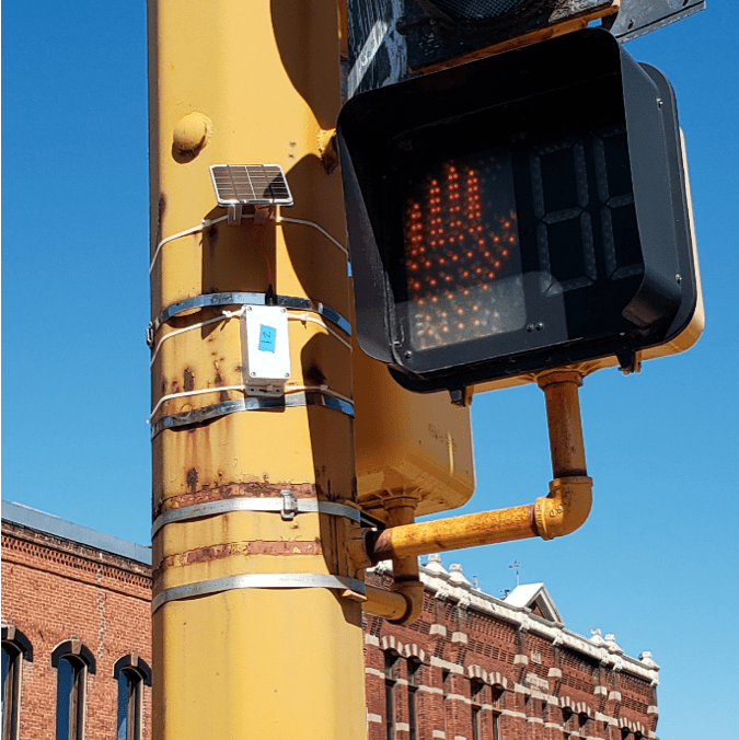 A small, white rectangular BLE beacon is attached to a traffic signal pole near the pedestrian signal light.