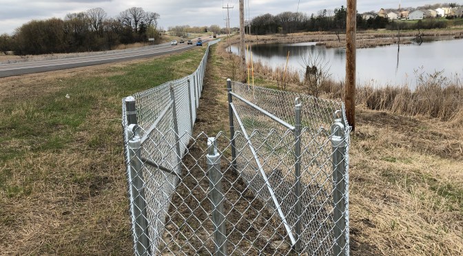 A chain-link fence, wrapping around in a “J” shape at the end, separates a roadway from a pond.