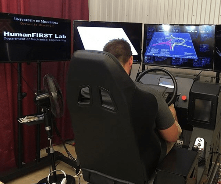 A person sitting in a chair looks at a series of computer screens attached to a console with steering wheel and shifter similar to a driver’s seat of a car. The main screen displays an aerial blueprint of an intersection.