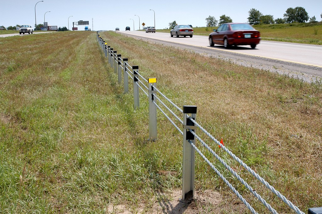 A cable barrier with three strands of cable in a grassy median next to a road.