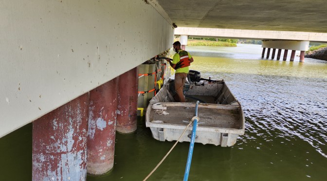 A worker in safety gear stands on a boat while repairing bridge piles.