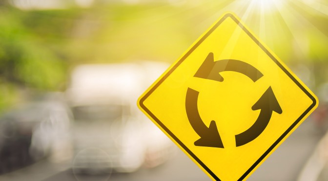 A yellow diamond-shaped roundabout sign with three arrows in a circle in the foreground with sunlight scattered and motor vehicles in soft focus in the background
