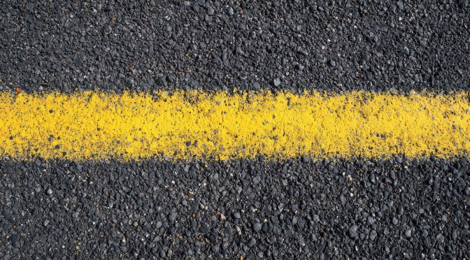 A shot of textured pavement and a yellow centerline stripe centered from above and somewhat close up.