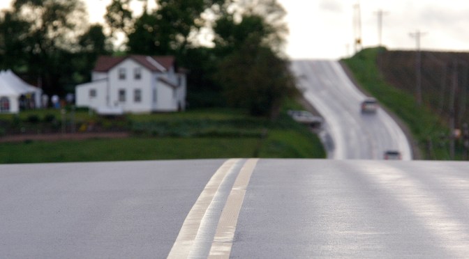 A hilly paved road shows a sheen of moisture on the surface as a car drives in the distance.