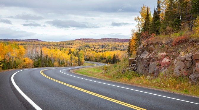 A winding new asphalt road disappears behind a rock bluff and into a forested Minnesota landscape in fall color.